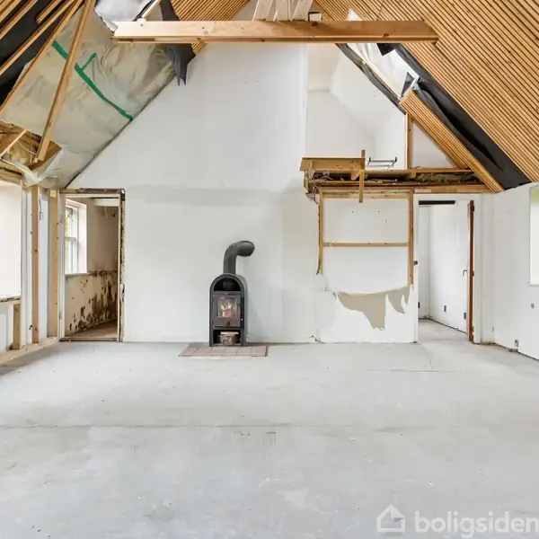 Et rummeligt rum med synlige loftsbjælker og en brændeovn centralt placeret; vægge er ubehandlede, og vinduer viser ud til en grøn have.