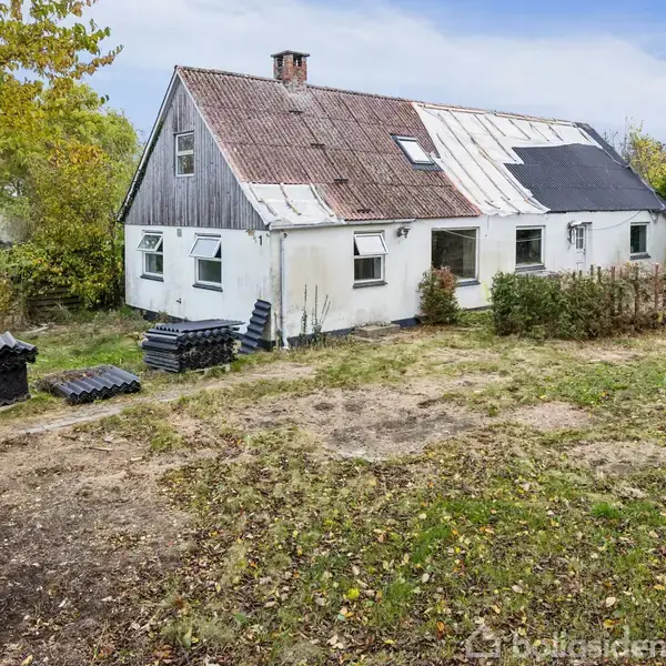 Et gammelt, hvidt hus med skråt tag i forskellige farvetoner står på en græsplæne. Stabler af tagsten er ved siden af huset, omgivet af træer og buske.