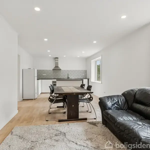 En sort lædersofa står i en moderne stue-køkken-alrum. I midten ses et træspisebord med stole. Rummet har hvide vægge, trægulv og store vinduer, der bringer lys ind.