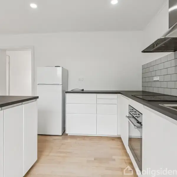 Et køkken med hvide skabe og sorte bordplader. Et komfur og emhætte er integreret. Køleskab står i hjørnet. Væggene er hvide med en grå flisebelægning. Ingen tekst til stede.