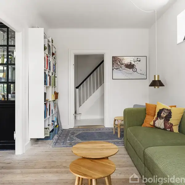 Grønt sofaarrangement med puder står i stue ved siden af en indbygget bogreol og et lille rundt bord. Et maleri hænger over sofaen. Rummet er lyst og åbent med trægulv.