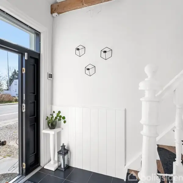 En lys entré indeholder en åben hoveddør, en sort lanterne og en plantestativ. Vægdekorationer af tre kuber pryder rummet. Sorte fliser på gulvet og en hvidmalet trappe leder opad.