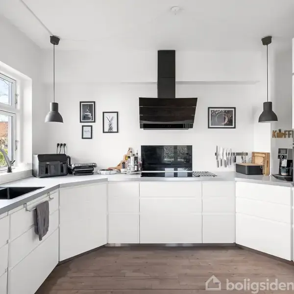 Hvidt, moderne køkken med sort emhætte over komfur. To hængelamper lyser over grå bordplader. Billeder hænger på væggen. Kaffemaskine ved siden af ovnen. Vindue til venstre.