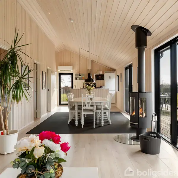 Stue med en spiseplads har et moderne brændeovn i hjørnet. Store glasdøre fører ud til en terrasse og have. Lyse vægge og gulve skaber en varm atmosfære.