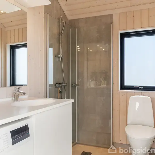 Badeværelse med en håndvaskenhed og vaskemaskine, bruser med glasvæg, og toilet. Væggene er beklædt med træpaneler, og et vindue giver lysindfald. Orden og minimalisme præger rummet.