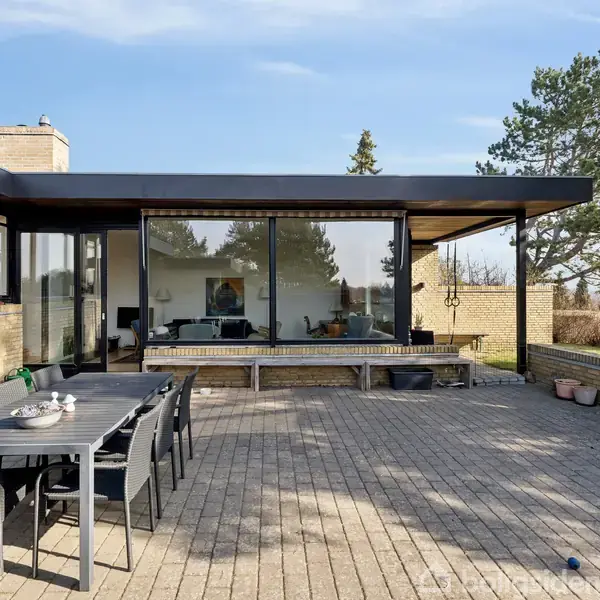 Et moderne hus med store glasfacader vender ud mod en flisebelagt terrasse. Sorte havemøbler står foran huset, omgivet af murstensvægge og træer i baggrunden.