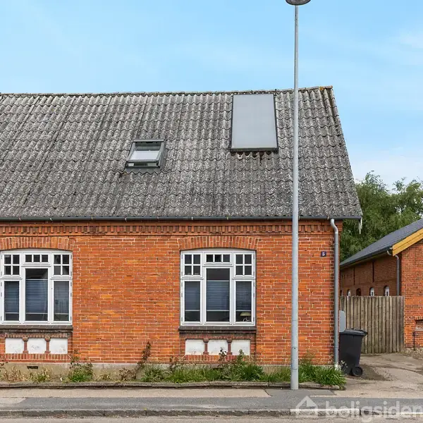 Et rødt murstenshus med to hvide vinduer og skråt tag ses fra vejen i en landsby. Til højre en tilstødende bygning og en skraldespand nær en lygtepæl.