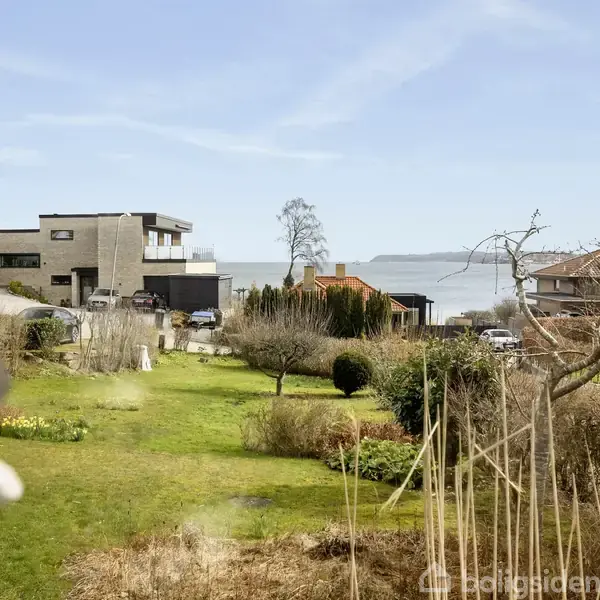 Moderne hus står roligt i villakvarter, omgivet af velplejede haver. I baggrunden ses havet under en klar blå himmel, hvilket skaber en fredfyldt atmosfære.