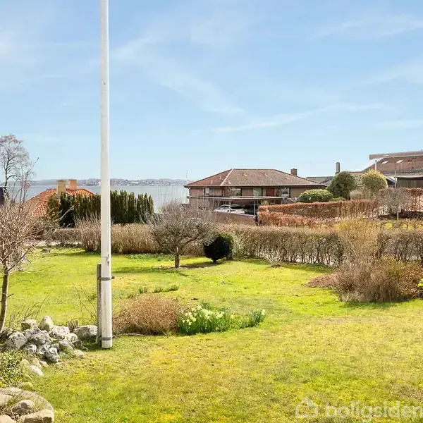Flagstang står i en grøn have med buske og træer, omkranset af hække. I baggrunden ses huse og en udsigt til vandet under en blå himmel.