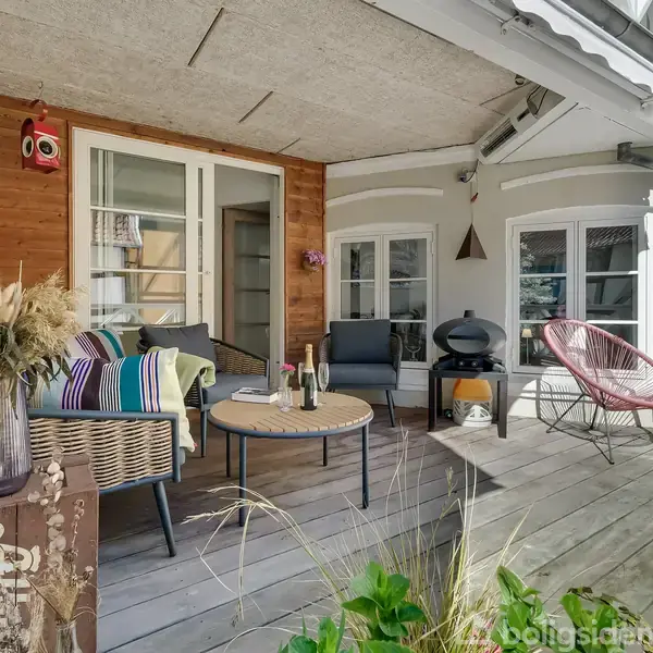 Terrasse med sofa og stole står omkring et stort rundt bord. Farverige puder og tæpper skaber en hyggelig atmosfære. Omkring er planter og et hvidt rækværk.