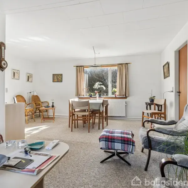 Stuen har en neutral indretning med lys sofa og lænestol, et tæppe på gulvet, et spisebord med stole, og et stort vindue med udsigt til haveplanter udenfor.