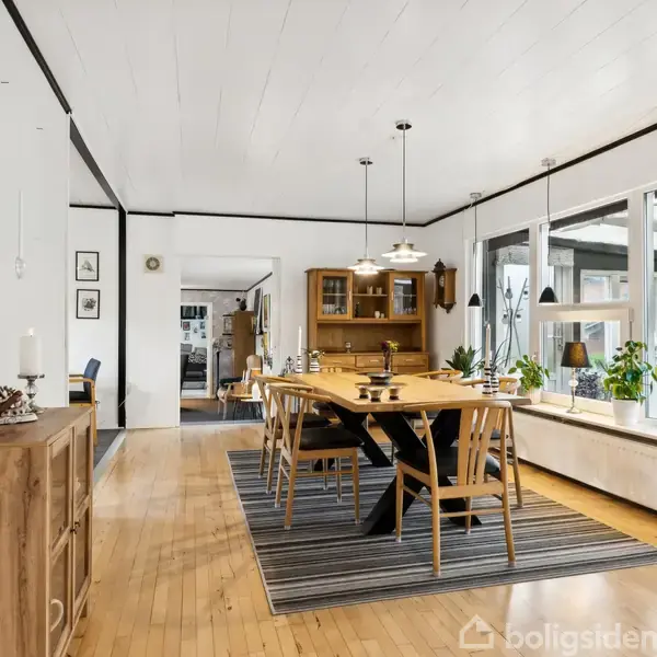 Spisebord med seks stole står på et stribet tæppe i en lys spisestue. Store vinduer fyldt med grønne planter omgiver rummet. En skænk står til venstre.