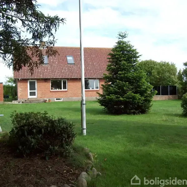 Et rødt murstenshus med skråt tag står i en grøn have med store træer og en flagstang. Til venstre står et bord-bænkesæt.