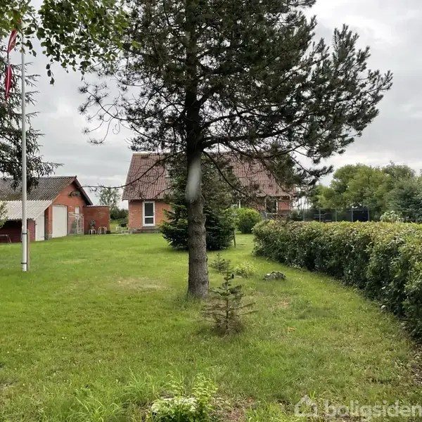 Et rødt murstenshus med tilhørende garage ses fra haven, hvor et flagstang står blandt grønne buske og træer. Skyer dækker himlen i baggrunden.