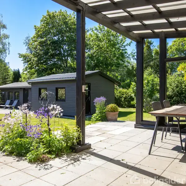 Terrasse med spisebord og stole står under en pergola. I haven ses blomsterbede, en grå havebygning og grønne træer, der skaber en rolig atmosfære.