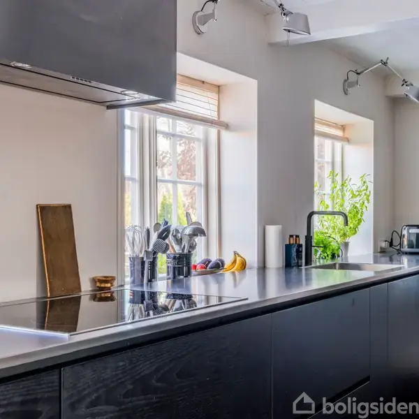 Flad sort køkkenbordplade med køkkenredskaber står under to store vinduer. Rustfrit emhætte hænger over bordet. Rummet er lyst og moderne indretning med planter i baggrunden.