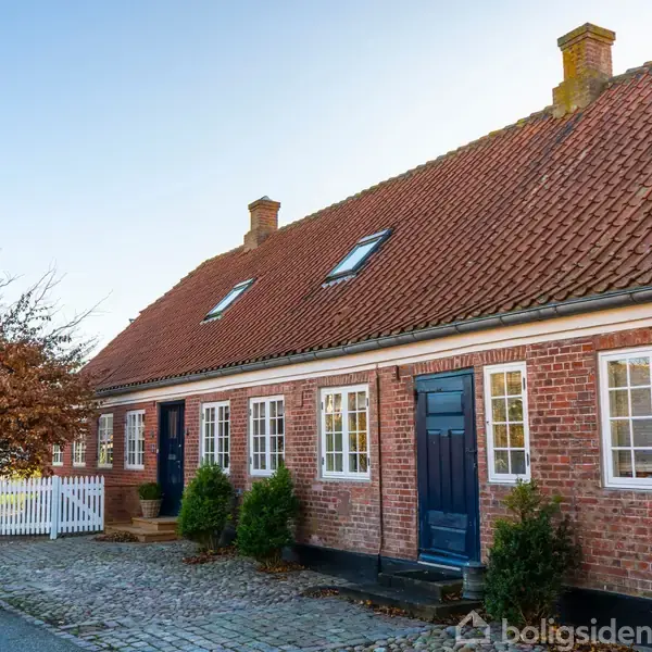 Rødstenshus med brunt tegltag har flere kviste og blå døre. Står ved brolagt vej og lille have med hvidt hegn. Træer uden blade i nærheden.