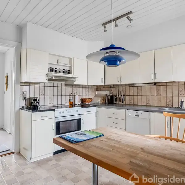 Et lyst køkken med hvide skabe og en sort bordplade. Et træbord med en stol står foran, og en blå lampe hænger over bordet. I baggrunden ses en dør til en anden del af huset.