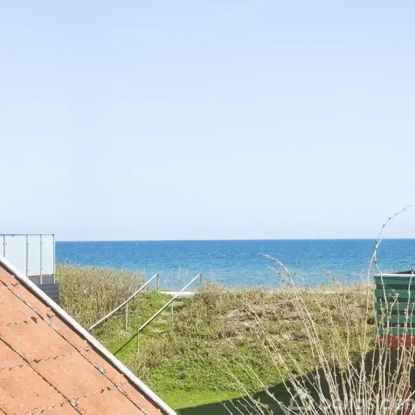 Rødt tag ligger mod venstre, med græs, en trampolin, og havet i baggrunden under en klar blå himmel.