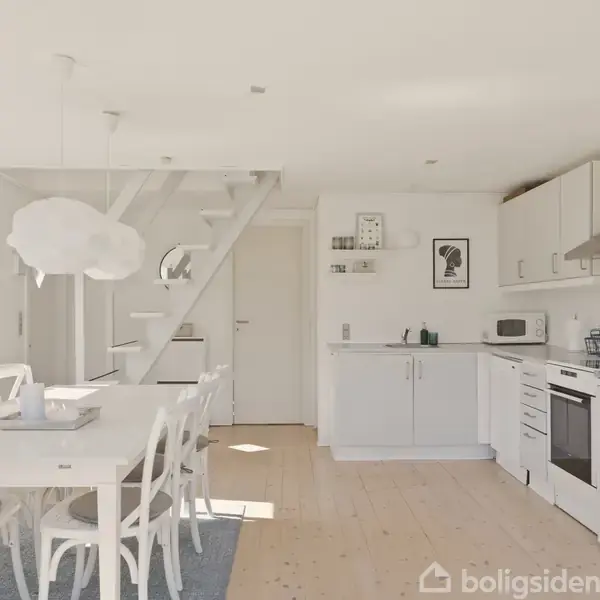 Køkken-alrum med hvidt spisebord, omkring syv stole, moderne køkkenindretning. En trappe fører opad. Stort vindue giver naturligt lys. Enkel og lys indretning med trægulv og hvidmalede vægge.