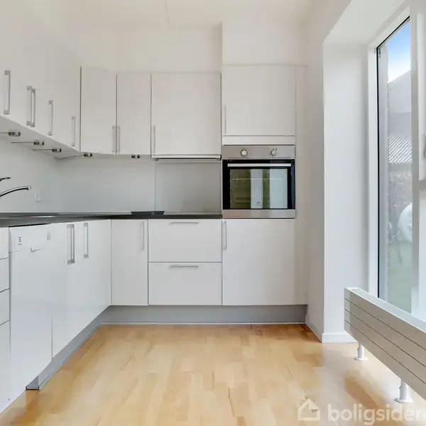 Et moderne køkken med hvide skabe og indbyggede ovne. Trægulv og store vinduer med udsigt til en have. Der er også en køkkenvask og køleskab.