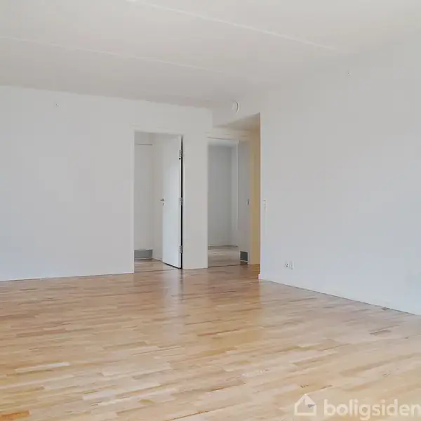 Et tomt rum med lyse trægulve og hvide vægge. En åben dør fører til en tilstødende gang. Rummet er lyst og enkelt i udtryk.