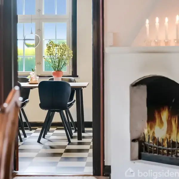 En pejs med levende ild står til højre, mens et spisebord med stole og en plante i en potte er synlig gennem en døråbning i baggrunden.