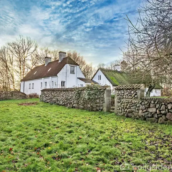 En hvidmalet landejendom står stille bag en stenmur, omgivet af frostbeklædte træer under en blå himmel. Græsset i forgrunden er grønt og frodigt.