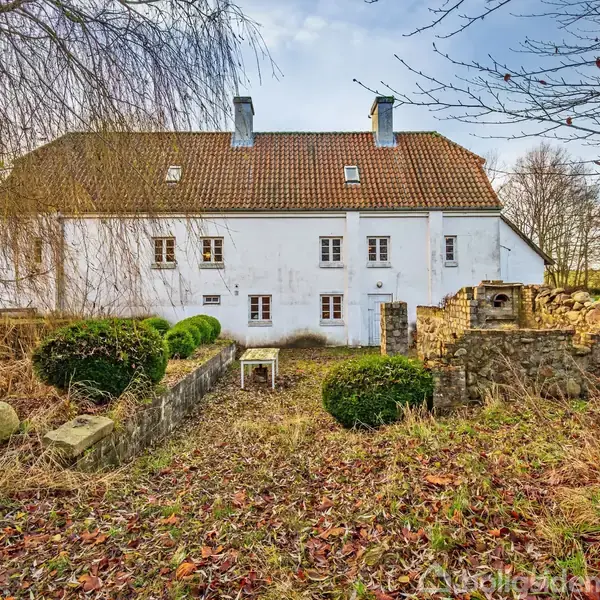 En hvid bygning med rødbrunt tegltag står stille i en landlig have med træer og buske omkring. Jorden er dækket af visne blade.