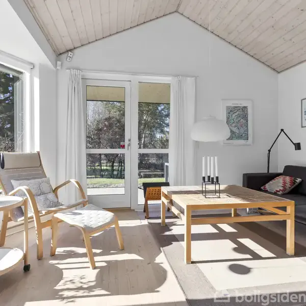 En stue med en sort sofa stående ved en væg med kunst. Et træbord med lys er foran sofaen. En lænestol ved et bord står tæt på vinduer, der giver udsigt til træer udenfor.