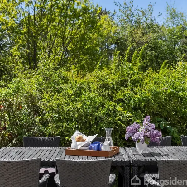 Terrassebord med flettede stole har en bakke med morgenmad og en vase med syrener. Sollys skinner gennem åbne døre mod grønne buske og træer i baggrunden.