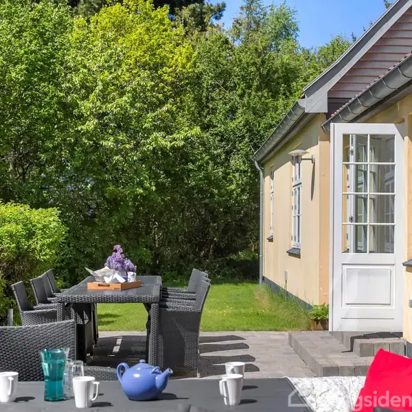 Et udendørs spisebord med stole står på en terrasse ved et gult hus. Bordet er dækket med kopper og en blå tekande. Omkring er der frodig vegetation.
