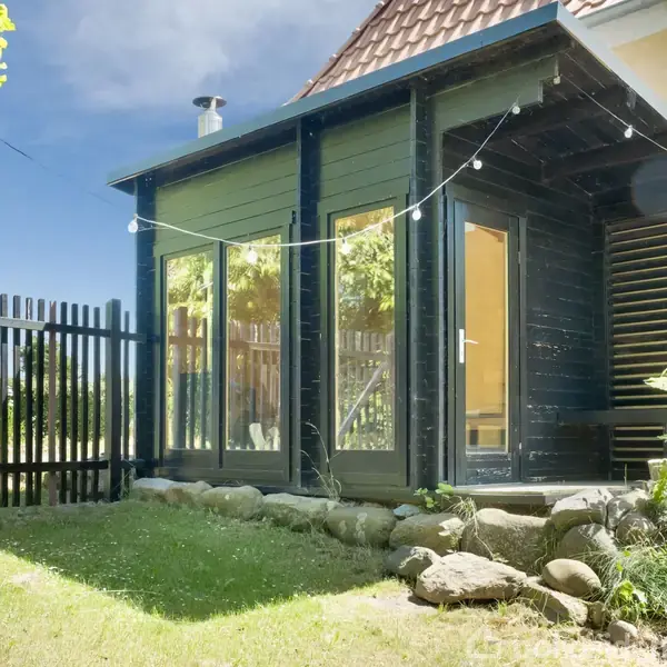 En lille, sort træbygning med store glasvinduer står ved siden af et gult hus. Der er lyskæder strakt over gårdspladsen og omgivet af en græsplæne og hegn.