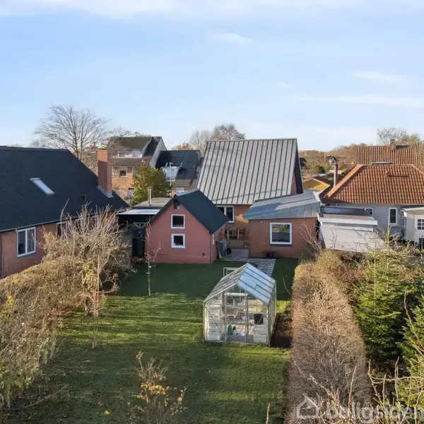Et drivhus står stille i en baghave blandt græs og buske. Baggrunden viser flere huse med forskellige tagtyper i en villakvarter.