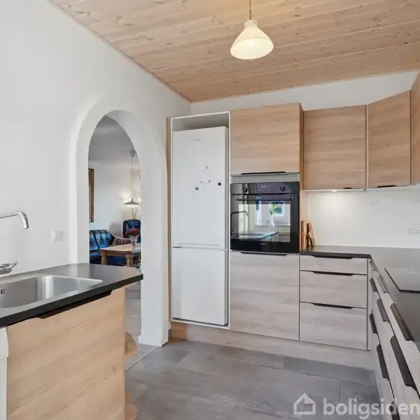 Køkken med træskabe og sort bordplade. Integreret ovn og køleskab. Rundbue fører til stuen med blå stole. Lysegråt gulv og træloft. Lampe hænger fra loftet.