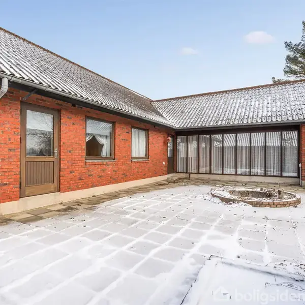 Rødt murstenshus med snedækket tag og indgangsparti, der fører til en stenterrasse. Et rundt bed er placeret centralt. Omgivet af høje træer og vinterlandskab.