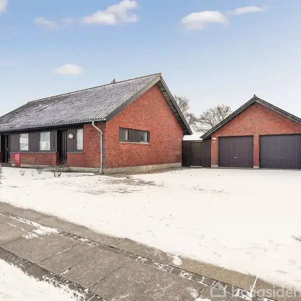 En rød murstenshus står stille ved siden af en to-garagebygning. Sne dækker jorden, og en grå fortovslinje løber foran dem. Himlen er let overskyet.