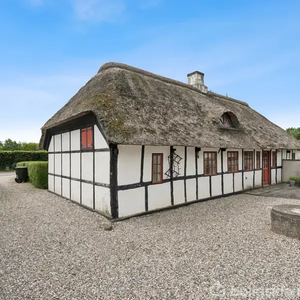 Bindingsværkshus med stråtag står i en gårdsplads med grusbelægning. Det er omgivet af hække og grønne træer under en blå himmel. Der er patio møbler udenfor.