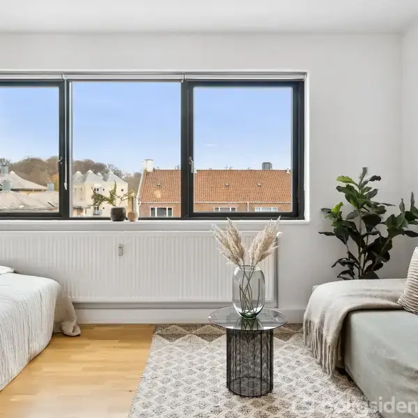 En seng står ved et stort vindue, der viser tage og træer udenfor. Ved siden af er en sofa med puder og et tæppe, samt et lille bord med planter i en vase.