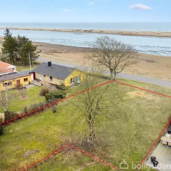 En gul villa står stille i en græsplæne med en rød markering omkring sig. I baggrunden ses en vej, træer og en flod.