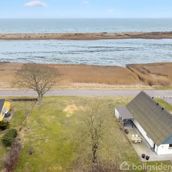 Hus med gråt tag og hvid facade ligger ved en vej, omgivet af græsplæne og et træ. I baggrunden ses en sø og havet.