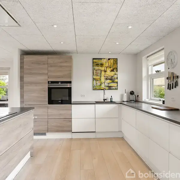 Et moderne køkken med lyse trægulve og hvid indretning. Elektriske apparater er indbygget i et skab, og vinduerne giver naturligt lys. En væg har et farverigt maleri og en ur.