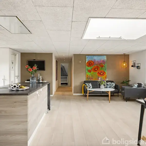 Køkkenø med frugtskål står i moderne, åbent rum med trægulv og lysindfald fra store vinduer. Loungeområde med sofa og farverigt maleri i baggrunden.