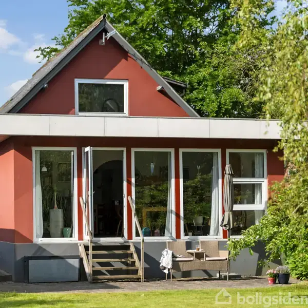 Et rødt hus med hvide døre og vinduer står i en grøn have. Trin fører til hoveddøren, og en terrasse har en parasol og stolarrangementer.