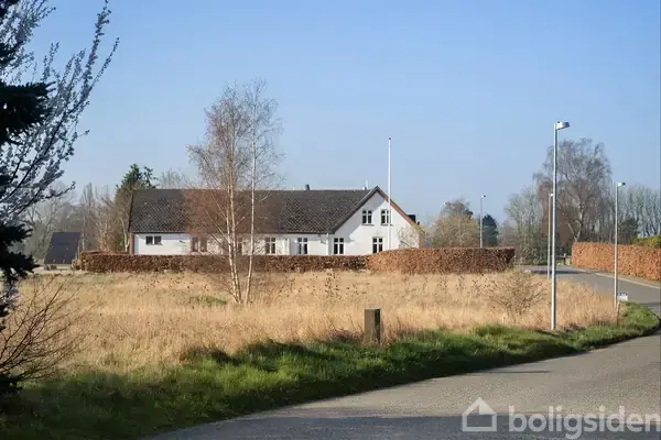 Hvidt hus ligger bag en lav hæk i en åben, græsbevokset område. En bøjelig vej og gadelygter markerer landskabet. Træer uden blade står rundt i det fjerne.