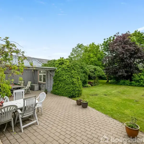 En havemøbelgruppe med hvide plastikstole og bord står på en flisebelagt terrasse. Omkring er der grøn beplantning og en velplejet græsplæne. I baggrunden ses et flagstang og buske.