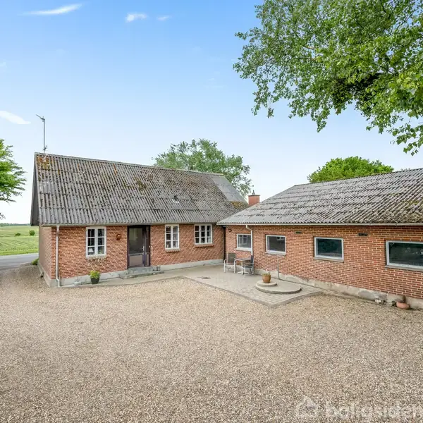 En rødstensbygning med to sektioner står på en grusbelagt plads med træer og grønt i baggrunden under en klar blå himmel.