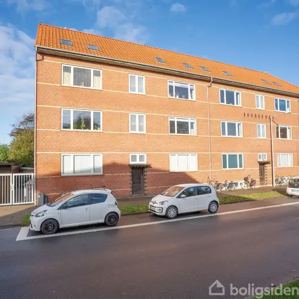 En tre-etagers murstensbygning med rødt tegltag står stille i et bymiljø. Foran holder flere biler parkeret langs fortovet på en asfalteret vej under en blå himmel.