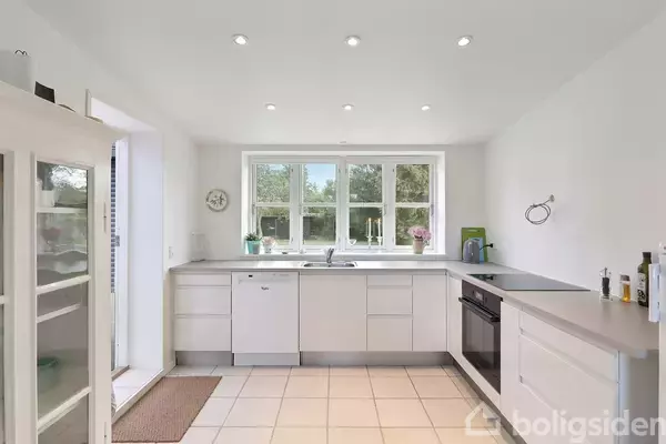 Hvidt køkken med moderne skabsindretning, opvaskemaskine og ovn. Stort vindue med udsigt til grøn natur. Omgivet af fliser på gulvet og spotbelysning i loftet.
