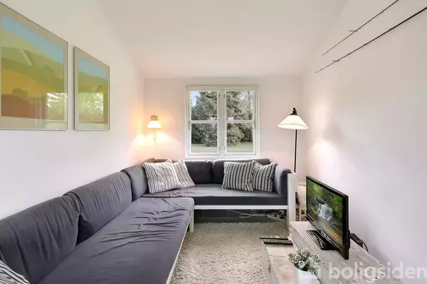 En grå sofa danner en hjørneformation, belyst af to lamper i en lys stue med hvidt tæppe. Et tv står på et lavt bord foran sofaen.